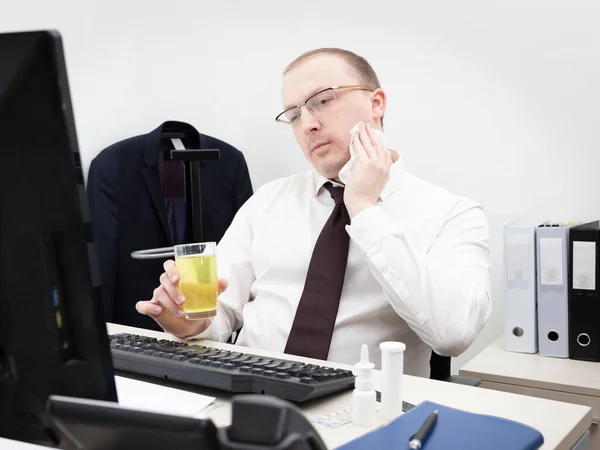 Sick man in a white business shirt and red tie working in office on a desktop computer, holds glass with medicine and wipes face with a napkin, close up