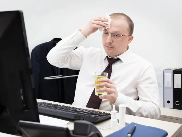 Sick man in a white business shirt and red tie working in office on a desktop computer, have got a headache, holds glass with medicine and wipes forehead with a napkin, close up