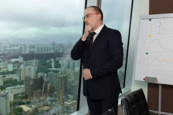Mavi takım elbiseli hoşnutsuz müdürün yan görünüş portresi ofis penceresinde yağmur ve bulutlara bakıyor, yakın çekim.