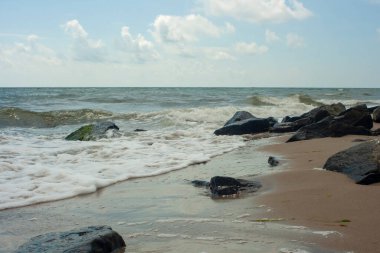 Köpüren deniz suyunun yatay fotoğrafçılığı. Deniz dalgaları, ıssız bir sahilde yeşil yosunlarla kaplı taşların üzerinde kırılıyor..