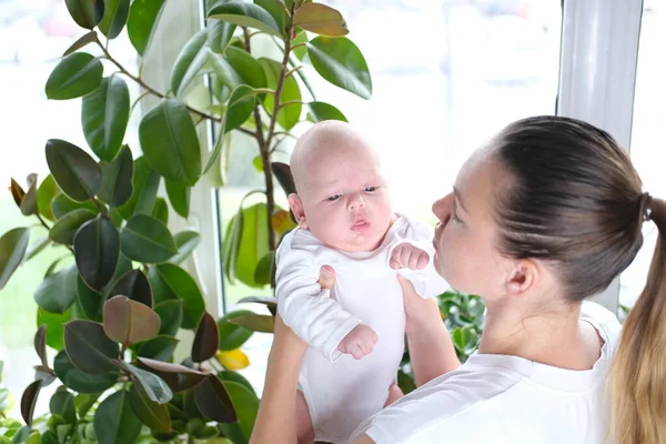 En vacker ung mor håller sin nyfödde son i famnen vid fönstret. Närbild av ett barn. — Stockfoto