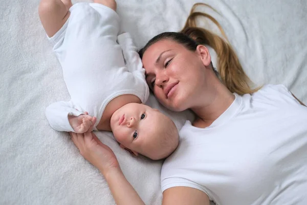 Bela jovem mãe está com seu bebê recém-nascido na cama.Retrato de mãe e filho em roupas brancas. — Fotografia de Stock