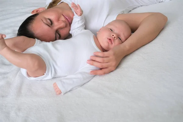 Bela jovem mãe está com seu bebê recém-nascido na cama.Retrato de mãe e filho em roupas brancas. — Fotografia de Stock