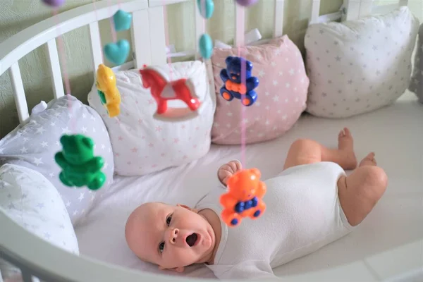 Pasgeboren baby in witte kleren ligt in de wieg en kijkt naar zijn speelgoed. De baby geeuwt close-up — Stockfoto