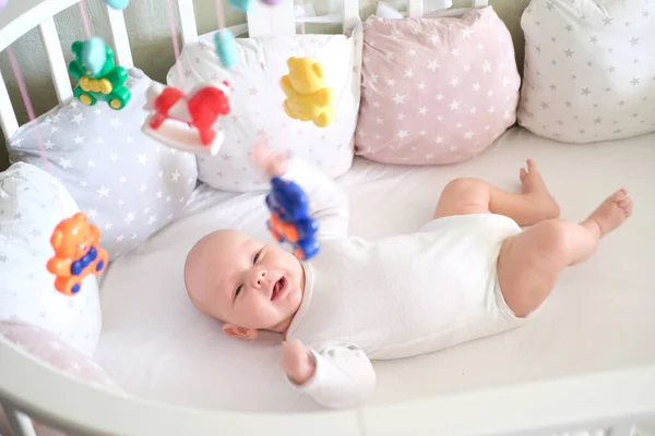Ett nyfött barn i vita kläder ligger i vaggan och tittar på sina leksaker. Baby leende på nära håll — Stockfoto