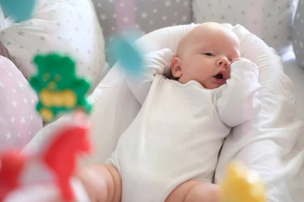 Ett nyfött barn i vita kläder ligger i vaggan och tittar på sina leksaker. Baby närbild — Stockfoto