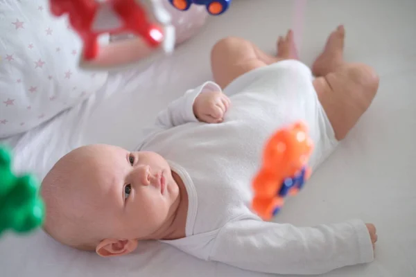 Ett nyfött barn i vita kläder ligger i spjälsängen och tittar på sina leksaker.Baby närbild — Stockfoto