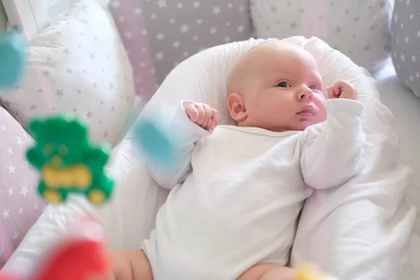 Новорожденный ребенок в белой одежде лежит в кроватке и смотрит на свои игрушки. Ребенок крупным планом — стоковое фото
