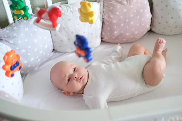 Ett nyfött barn i vita kläder ligger i vaggan och tittar på sina leksaker. Baby leende på nära håll — Stockfoto