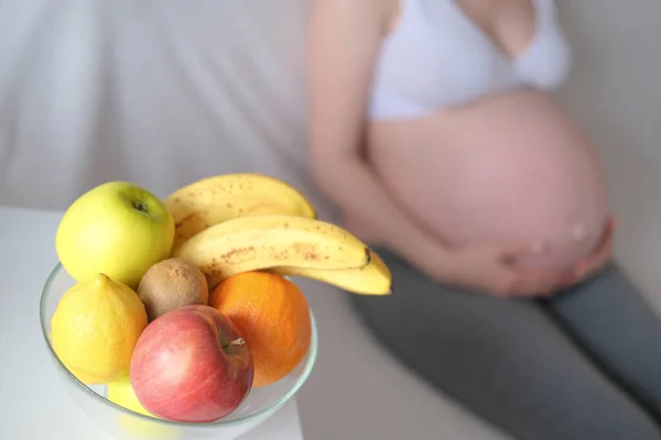 Alimentação saudável, conceito de gravidez. Barriga de uma mulher grávida em um fundo branco e um prato com frutas na mesa — Fotografia de Stock