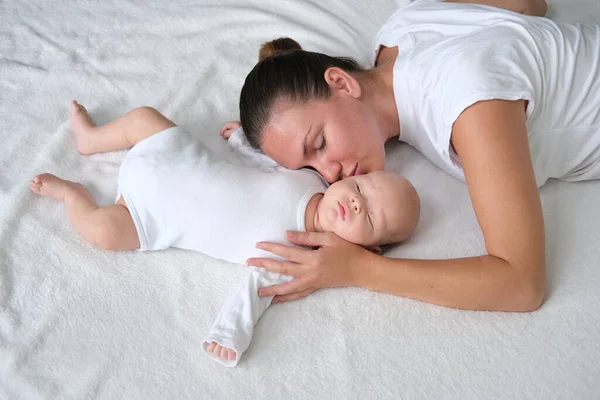 Bela jovem mãe está com seu bebê recém-nascido na cama.Retrato de mãe e filho em roupas brancas. — Fotografia de Stock