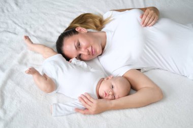 Güzel genç anne yeni doğmuş bebeğiyle yatakta yatıyor. Beyaz elbiseli anne ve oğlunun portresi..