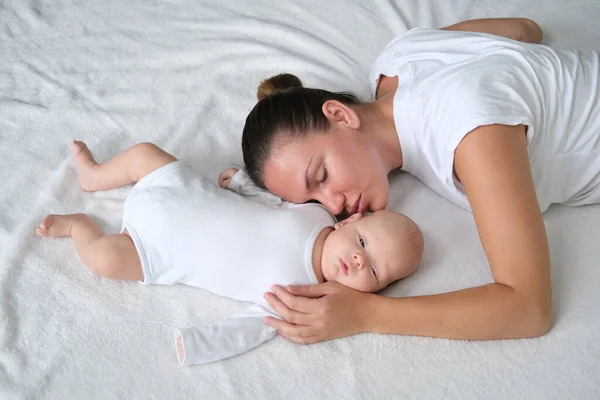 Hermosa madre joven se acuesta con su bebé recién nacido en la cama.Retrato de mamá e hijo en ropa blanca. — Foto de Stock