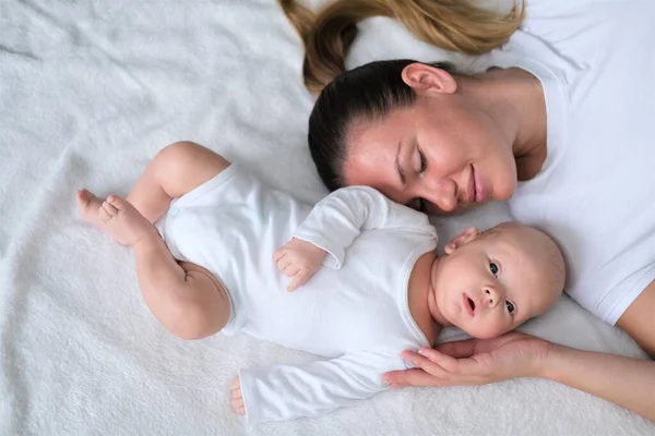 Bela jovem mãe está com seu bebê recém-nascido na cama.Retrato de mãe e filho em roupas brancas. — Fotografia de Stock