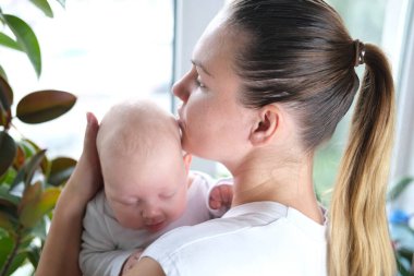 Güzel, genç bir anne yeni doğmuş oğlunu pencerenin kenarında kollarında tutuyor. Bir çocuğun yakın plan portresi.