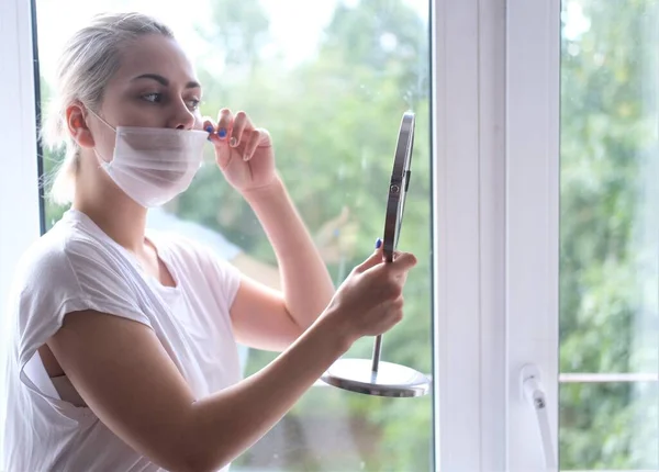 Young beautiful woman in a medical mask stands by the window.Portrait of a girl in a protective mask. — Stock Photo, Image