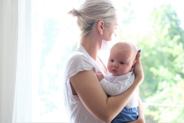 Koruyucu maskeli genç bir kadın yeni doğmuş bir bebeği pencerede tutuyor. Güzel bir genç kadın ve çocuğun portresi..