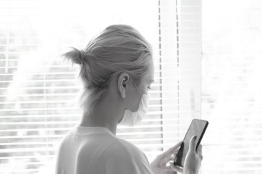 Tıbbi maskeli genç ve güzel bir kadın pencerenin yanında duruyor ve telefonda konuşuyor. COVID-19 'un Önlenmesi.