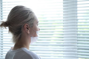 Tıbbi maskeli genç bir kadın pencerenin yanında duruyor. Koruyucu maskeli bir kızın portresi.
