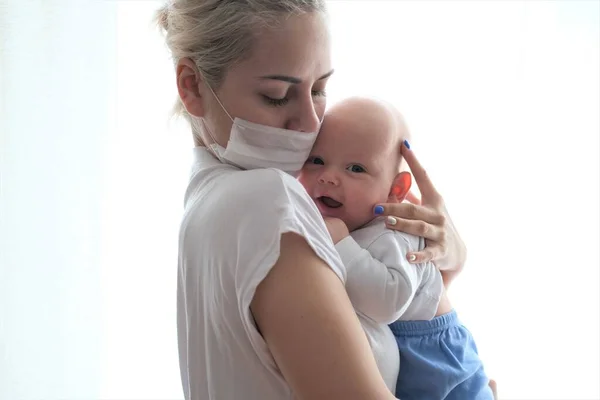 Koruyucu maskeli genç bir kadın yeni doğmuş bir bebeği pencerede tutuyor. Güzel bir genç kadın ve çocuğun portresi..