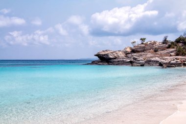 Yumuşak deniz dalgaları, beyaz kumlu tropikal bir sahil ve arka planda kayalar..