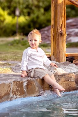 Nehir kıyısında küçük bir çocuk oturuyor. Suya bak. Bacaklarını suda sallıyor. Doğada yürümekten zevk alır..