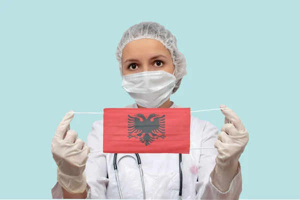 Portrait of a young woman doctor in a protective suit who holds in her hands medical protective mask with the image of the flag of Albania. Albania epidemic outbreak concept