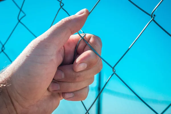 A man\'s hand holding on to the mesh netting or chain-link fencing against the background of a clear blue sky.