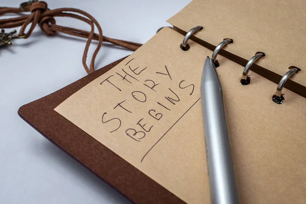 Blank notebook in a leather case, on the page with a pen it is written: The story begins. The concept of a new story, story, adventure, new page in life