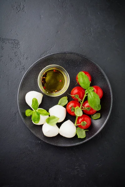 Kiraz domates, fesleğen, mozzarella peyniri ve zeytinyağı koyu arka plan üzerinde siyah plaka. İtalyan yemeği arka plan. Seçici odak. Üstten görünüm.