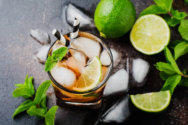 Summer cooling alcoholic beverage from cola, whiskey, lime and liqueur in glass on dark concrete old background.