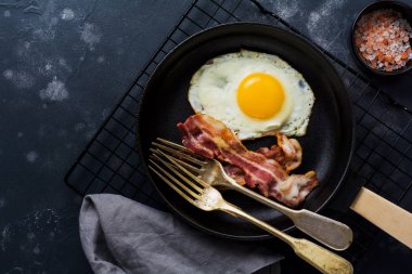 Geleneksel İngiliz kahvaltısı kızarmış yumurta ve pastırma ile koyu beton arka planda dökme demir tavada. Üst görünüm.
