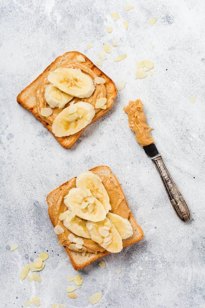 Toast with peanut butter, banana slices, honey and almond flakes on old gray concrete background. Top view.