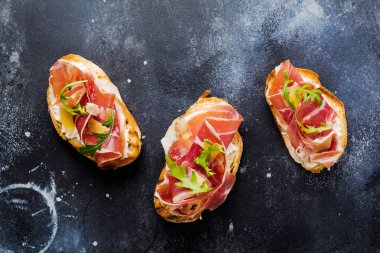 Jamon, roka ve beton eski koyu arka plan üzerinde sert peynir ile açık sandviçler. Rustik tarzı. Üstten Görünüm.