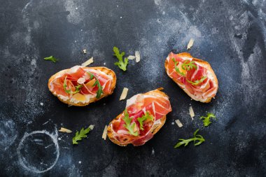 Jamon, roka ve beton eski koyu arka plan üzerinde sert peynir ile açık sandviçler. Rustik tarzı. Üstten Görünüm.