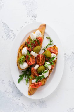 Beyaz tabak eski ışık arka plan üzerinde kiraz domates, mozzarella, zeytin ve fesleğen ile Caprese bruschetta tost. Üstte görüntü.