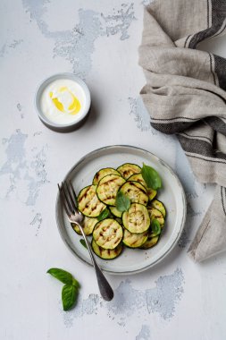 Beyaz beton arka plan üzerinde basit bir seramik tabakta fesleğen yaprakları ile ızgara kabak salatası. Düz yatıyordu