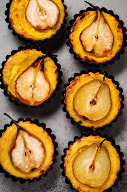 Koyu gri beton bir masa arkasında armut ve kremalı mini tartlar. Üst görünüm.