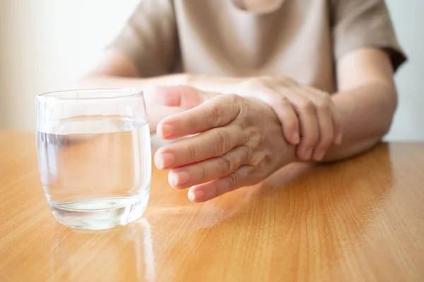 Elderly woman hands w/ tremor symptom reaching out for a glass of water on wood table. Cause of hands shaking include Parkinson's disease, stroke or brain injury. Mental health neurological disorder.