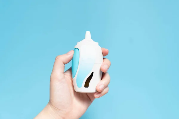 Hand holding steroid nasal spray suspension w/ blank label on light blue background. Medical device used to treat allergic rhinitis, hay fever, sinusitis or nasal polyps. Health care concept. Closeup.