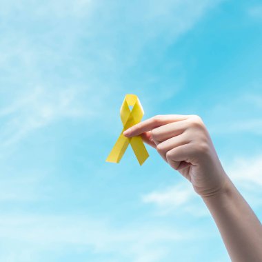 Mavi gökyüzünde sarı altın kurdeleyi tutan el. İntihar önleme, endometriyozis, sarkom kemik kanseri, mesane kanseri, karaciğer kanseri ve çocukluk kanseri farkındalığı konseptini destekleyen sembol.