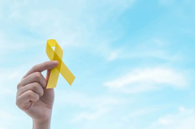 Mavi gökyüzünde sarı altın kurdeleyi tutan el. İntihar önleme, endometriyozis, sarkom kemik kanseri, mesane kanseri, karaciğer kanseri ve çocukluk kanseri farkındalığı konseptini destekleyen sembol.