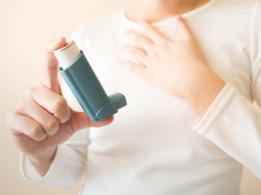 Young female in white t-shirt using blue asthma inhaler for relief asthma attack. Pharmaceutical products is used to prevent and treat wheezing and shortness of breath caused asthma or COPD. Close up. clipart