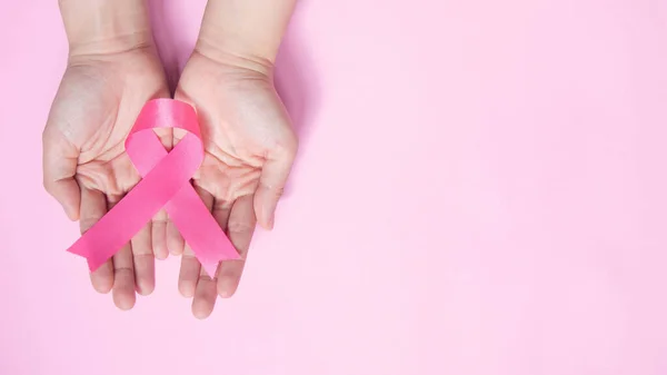 International symbol of Breast Cancer Awareness Month in October. Close up of female hand holding satin pink ribbon awareness on pink background w/ copy space. Women\'s health care and medical concept.