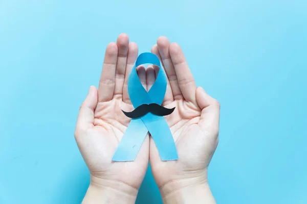 Men\'s health and Prostate cancer awareness campaign in November. Man hands holding light blue ribbon awareness w/ mustache on blue background. Symbol for support men who living w/ cancer.