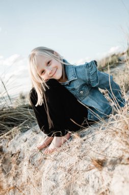 Kumsalda kumulda oturan genç bir kız kot ceket giymiş kameraya bakıyor.