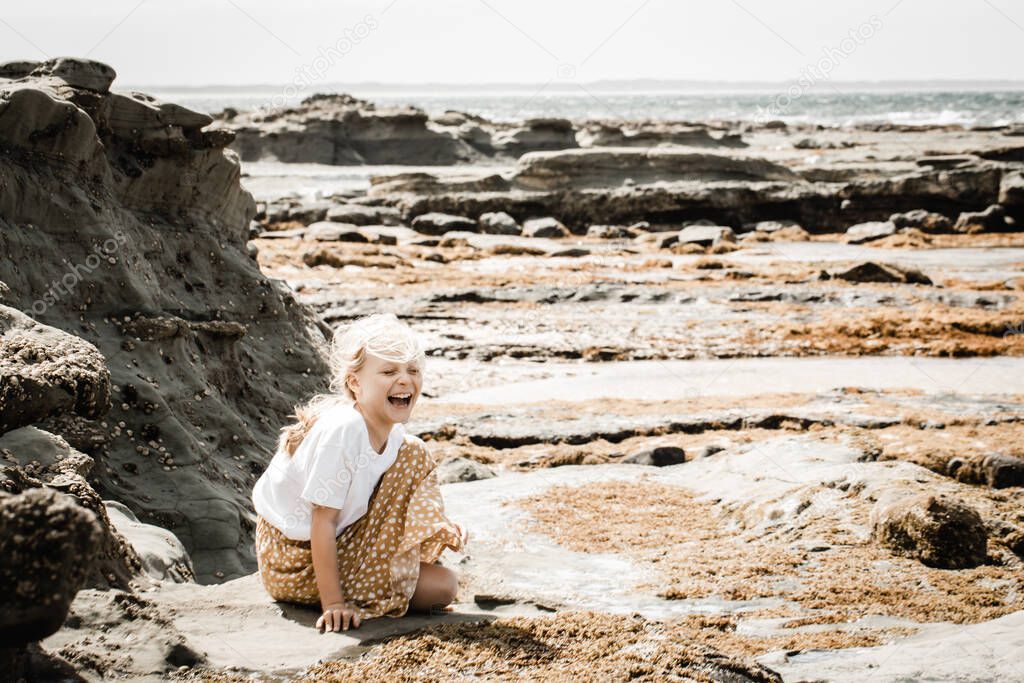 Young girl sitting looking at rockpools at the beach, there is cliffs with barnicles and seaweed aroung her, she is laughing