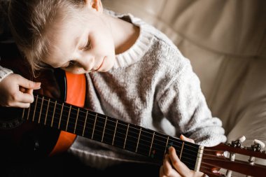 Pencerenin kenarında oturan, akustik gitar çalma alıştırması yapan sarışın bir kız.