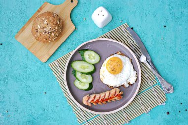 Kahvaltı, sosisli sahanda yumurta ve turkuaz beton arka planda kahverengi kil tabakta salatalık dilimleri. Çocuk kahvaltıları. Okul kahvaltı konsepti. Üst görünüm, alanı kopyala.
