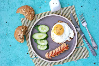 Kahvaltı, sosisli sahanda yumurta ve turkuaz beton arka planda kahverengi kil tabakta salatalık dilimleri. Çocuk kahvaltıları. Okul kahvaltı konsepti. Üst görünüm, alanı kopyala.